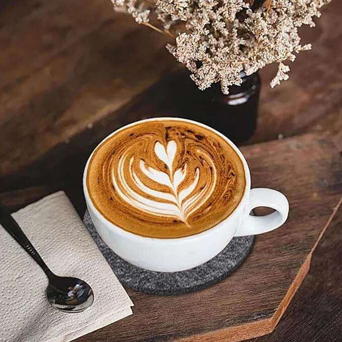Filzuntersetzer unter Tasse mit Latte Art neben einer Blume auf einem Holztisch.